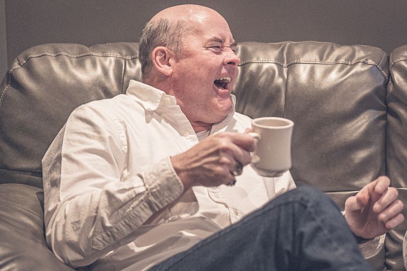 Man laughing out loud while holding a mug on a leather couch