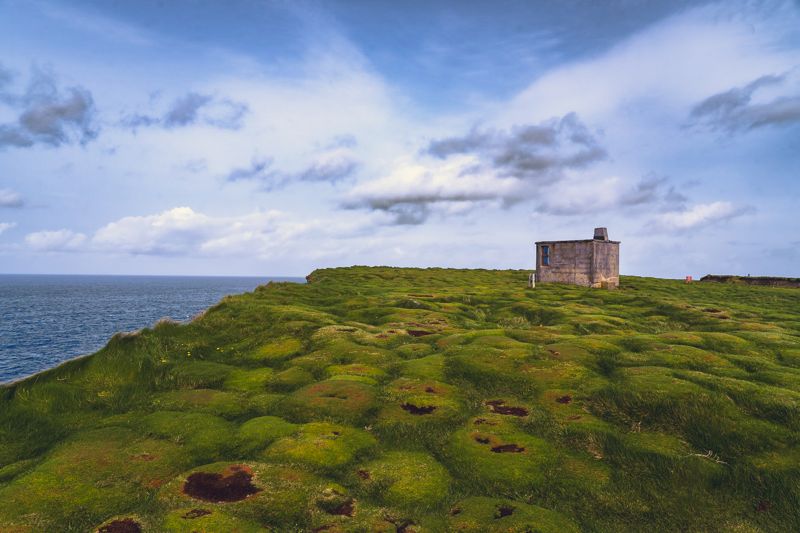 Stone hut on a green clifftop overlooking the sea under a cloudy sky