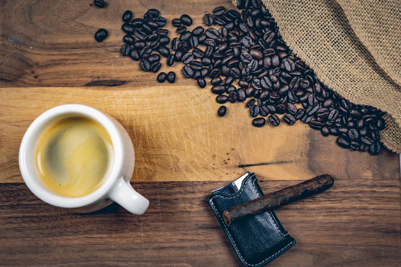 Overhead view of an espresso cup, scattered coffee beans, and a cigar on a wooden board