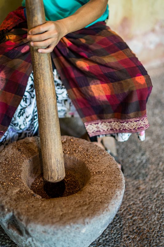 Hands holding a wooden pestle over a stone mortar filled with ground coffee