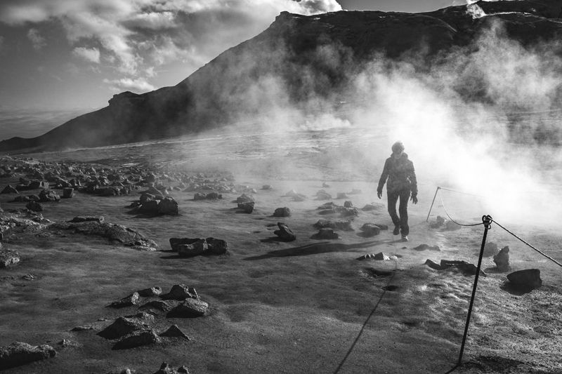 Black and white photo of a person walking through geothermal steam below a mountain