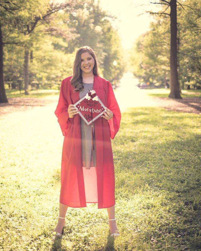 Graduate in a red gown holding her decorated cap in a tree-lined park