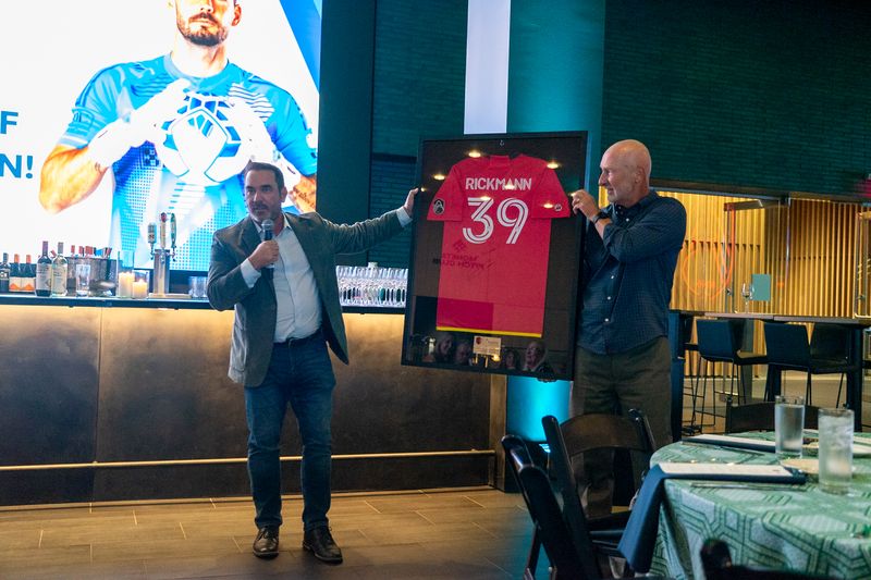 Two men holding a framed red jersey on stage at an evening event