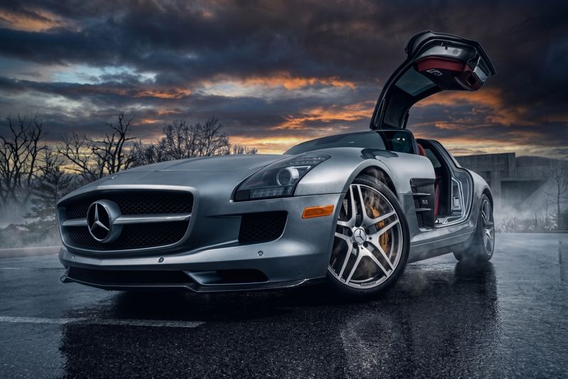 Silver sports coupe with its gullwing door raised on wet pavement under a dramatic sunset sky