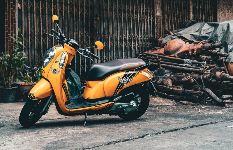 Yellow scooter parked in front of a rusted folding gate beside a pile of old machinery